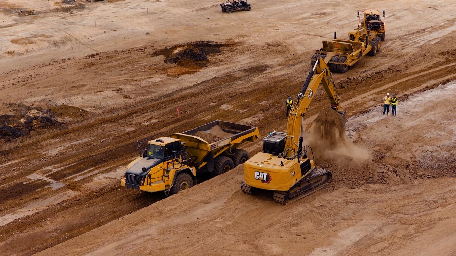 Aerial view of a CAT excavator loading dirt into a dump truck on a construction site, with workers and heavy equipment nearby.